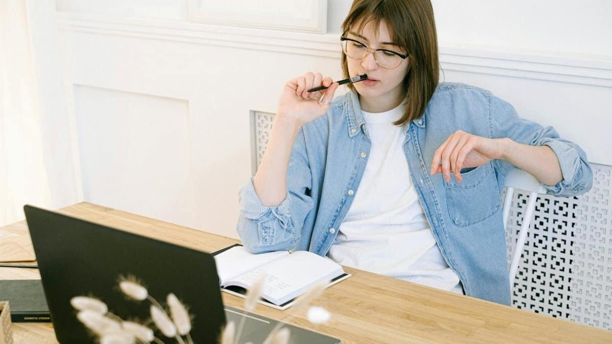 Woman working thoughtfully in home office, reflecting on ideas with laptop and notebook.