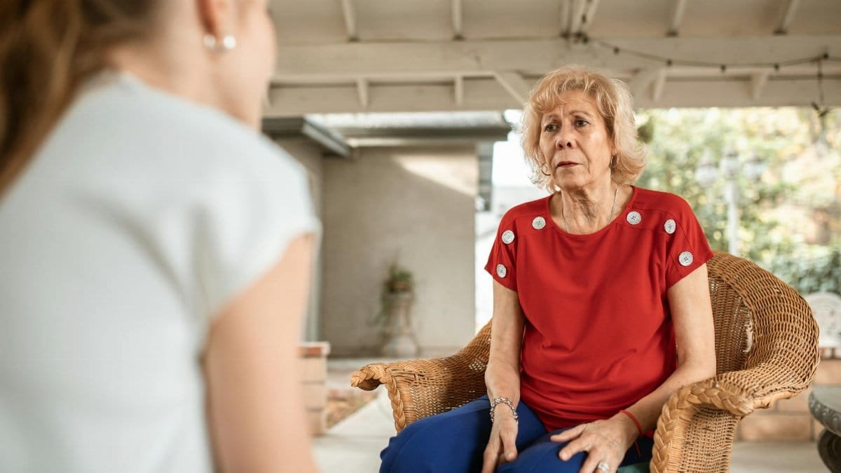 Senior woman in red shirt conversing indoors with younger woman. Daytime scene.