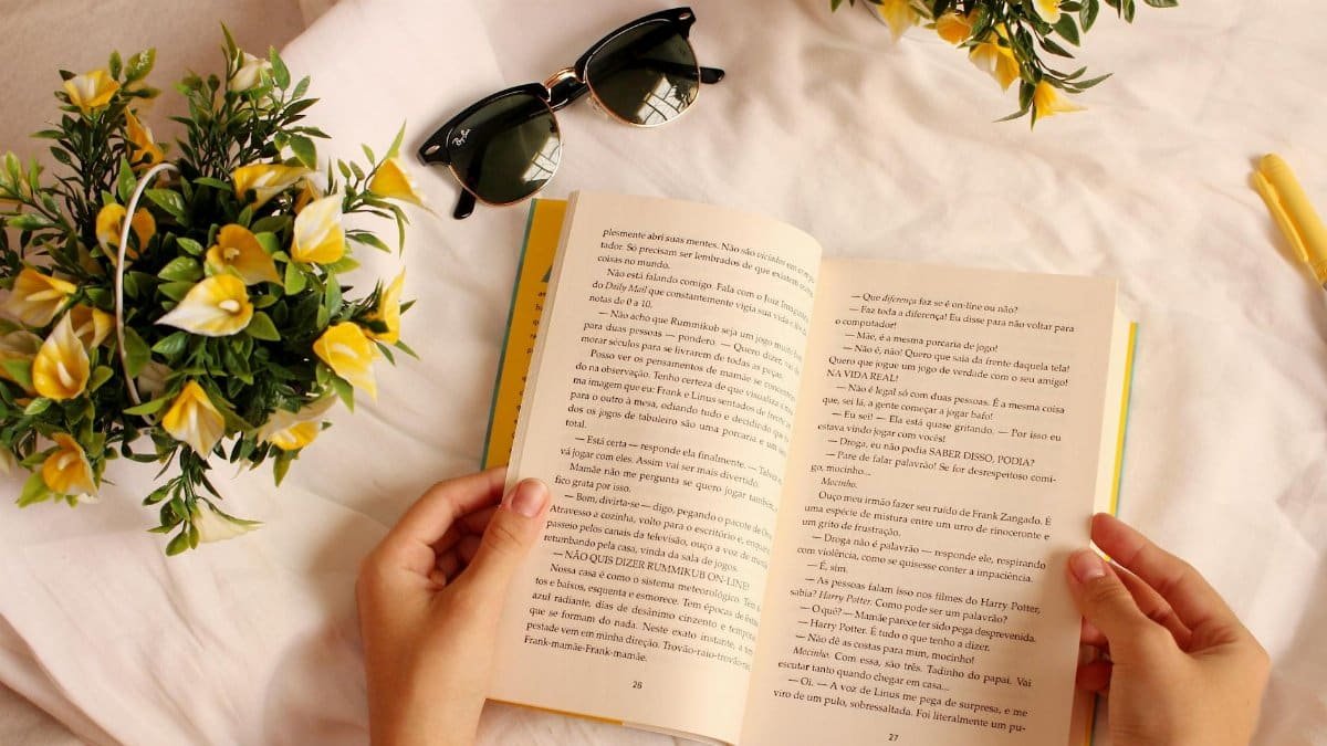 Hands holding a book surrounded by yellow flowers and sunglasses on a sunny day.