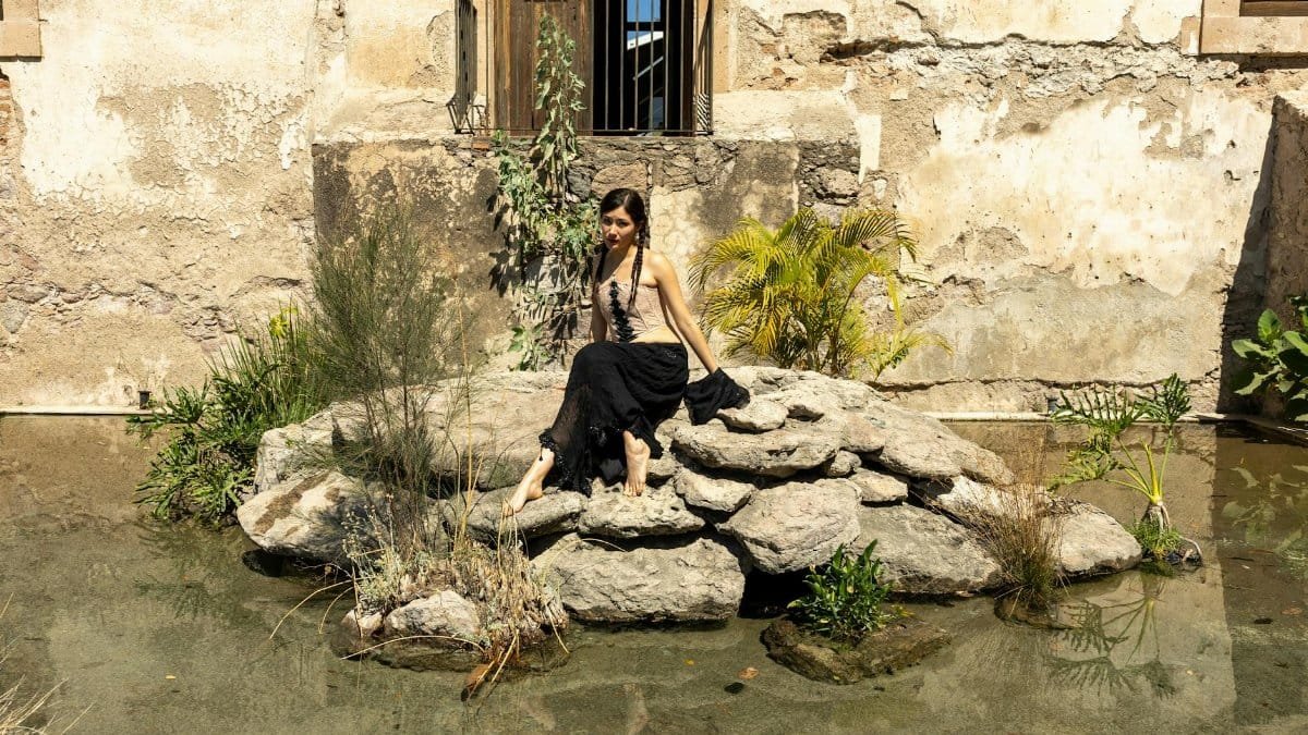 A woman rests on rocks near water outside a weathered building, showcasing rustic ambiance.