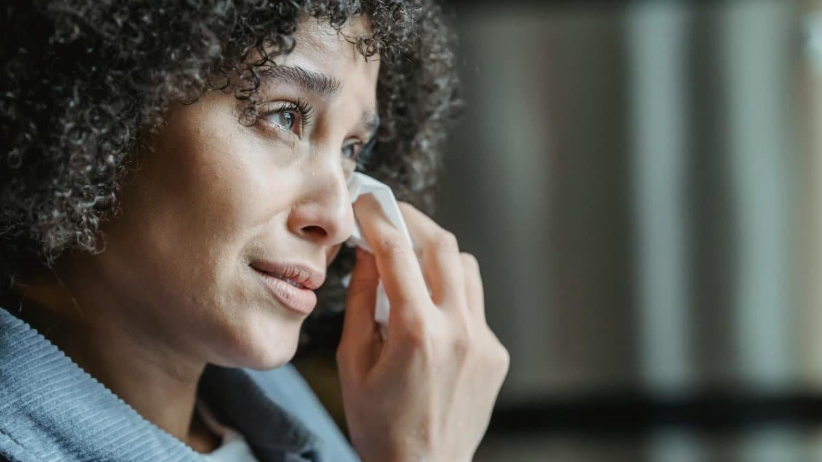 African American woman with curly hair wipes tears with tissue in a quiet indoor setting.