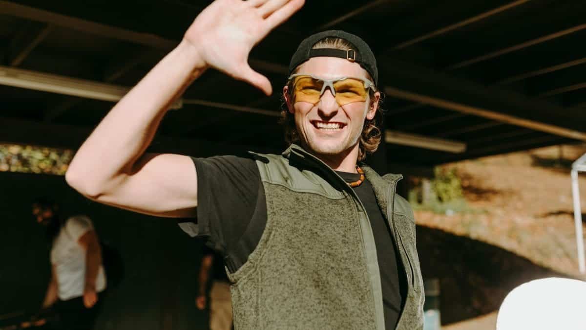 Smiling man wearing sunglasses and waving under a shaded structure outdoors.