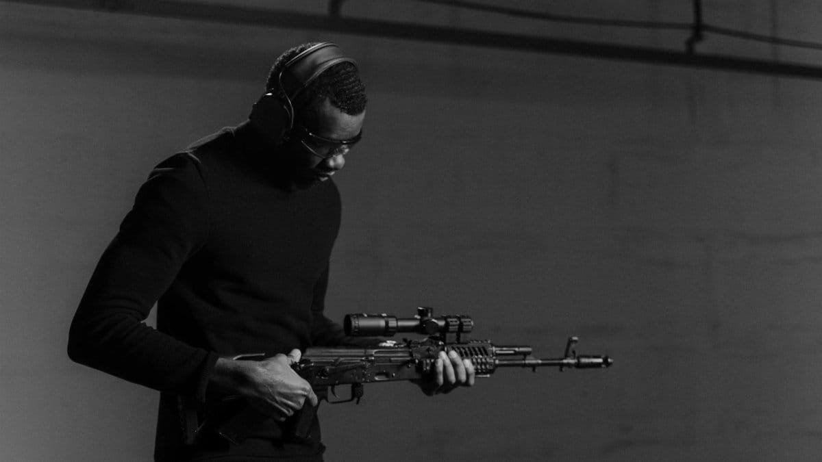Black and white image of a man holding a rifle, wearing headphones and eye protection.