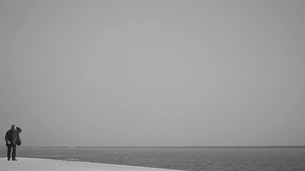 A solitary man stands on the shore, gazing at the vast, empty sea horizon.