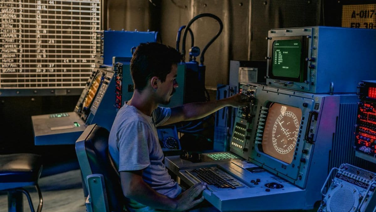 A man working at control panels in a dimly lit industrial room with various monitors.