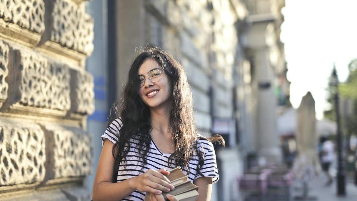 Happy young woman standing outdoors holding books, smiling in city setting.