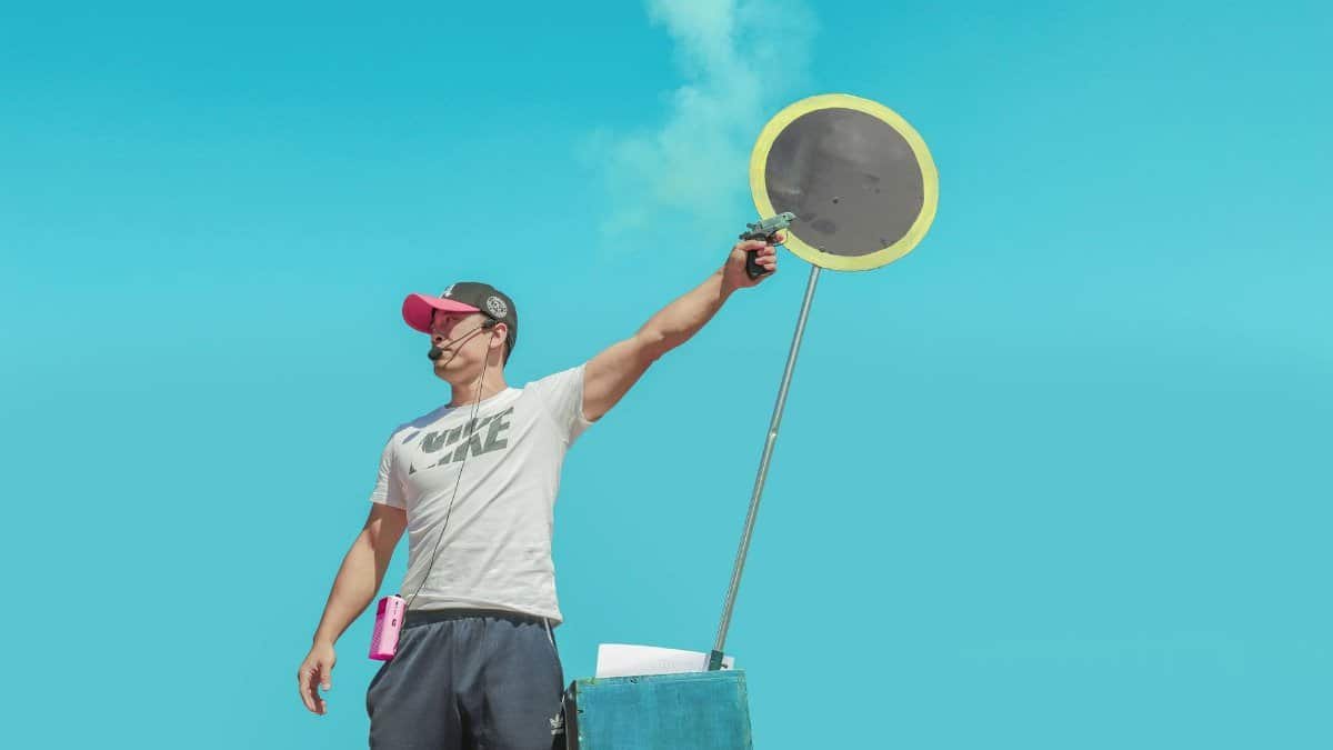 Man in cap and t-shirt with microphone signals race start against clear sky.
