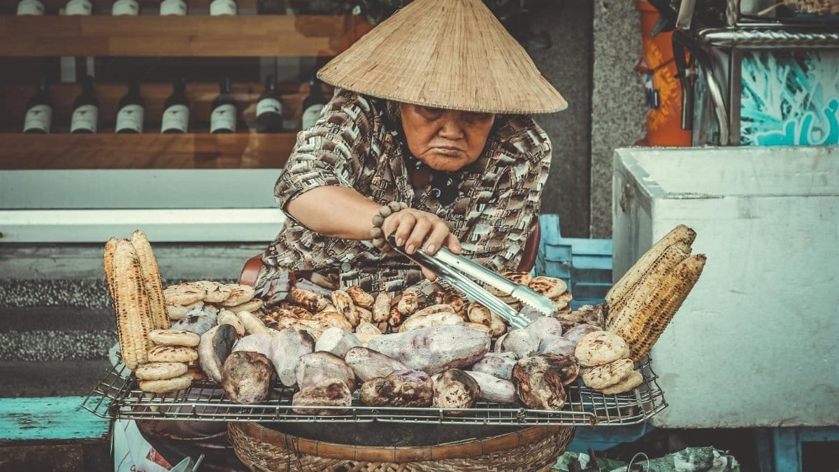 Asian woman grilling corn and sweet potatoes on a street food stand with a traditional hat.