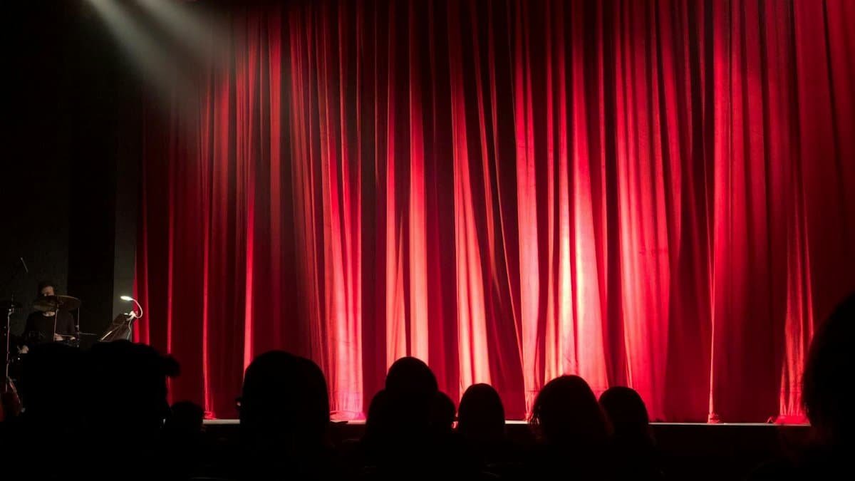 Dimly lit theater stage with red curtains and audience silhouettes under spotlights.