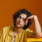 Young man with curly hair in vibrant yellow attire resting on a vintage TV set.
