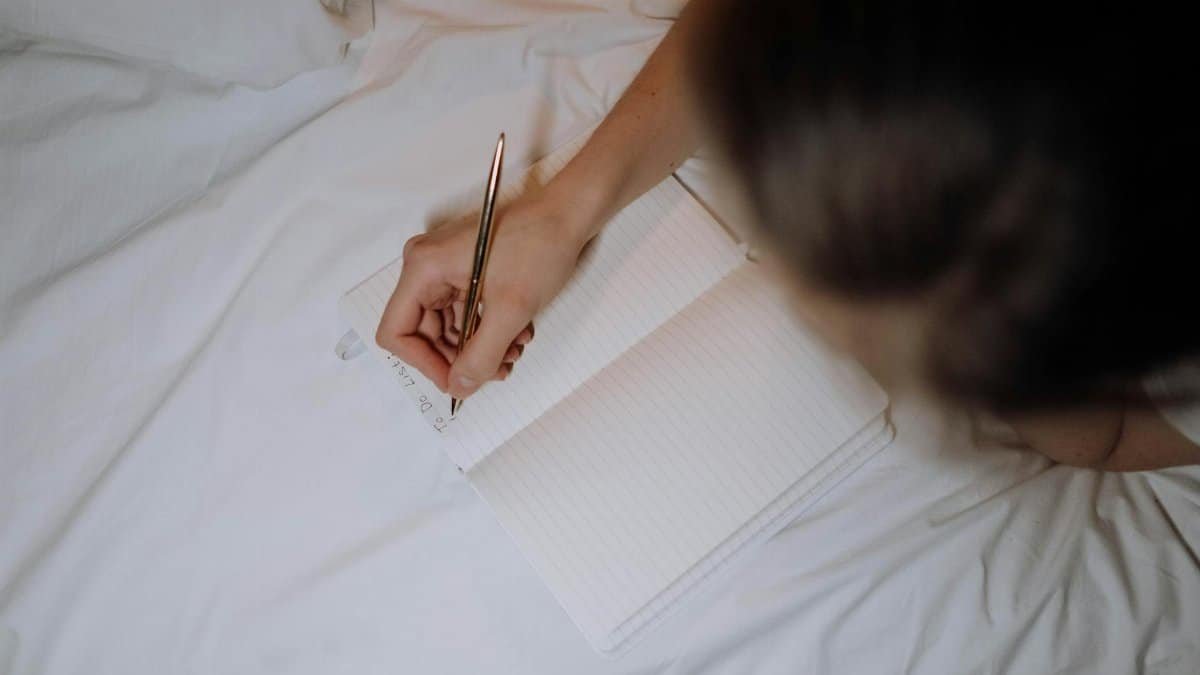 A person writing a to-do list in a notebook while sitting on a bed with white linen.
