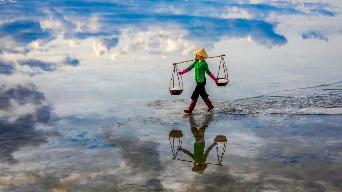 A worker carrying salt in baskets walks across a reflective water surface under a cloudy sky.