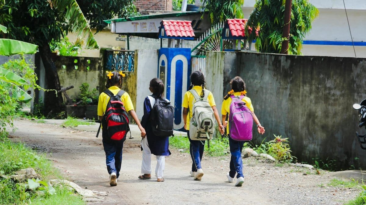 Four school children walk on an unpaved road, carrying backpacks, surrounded by greenery.