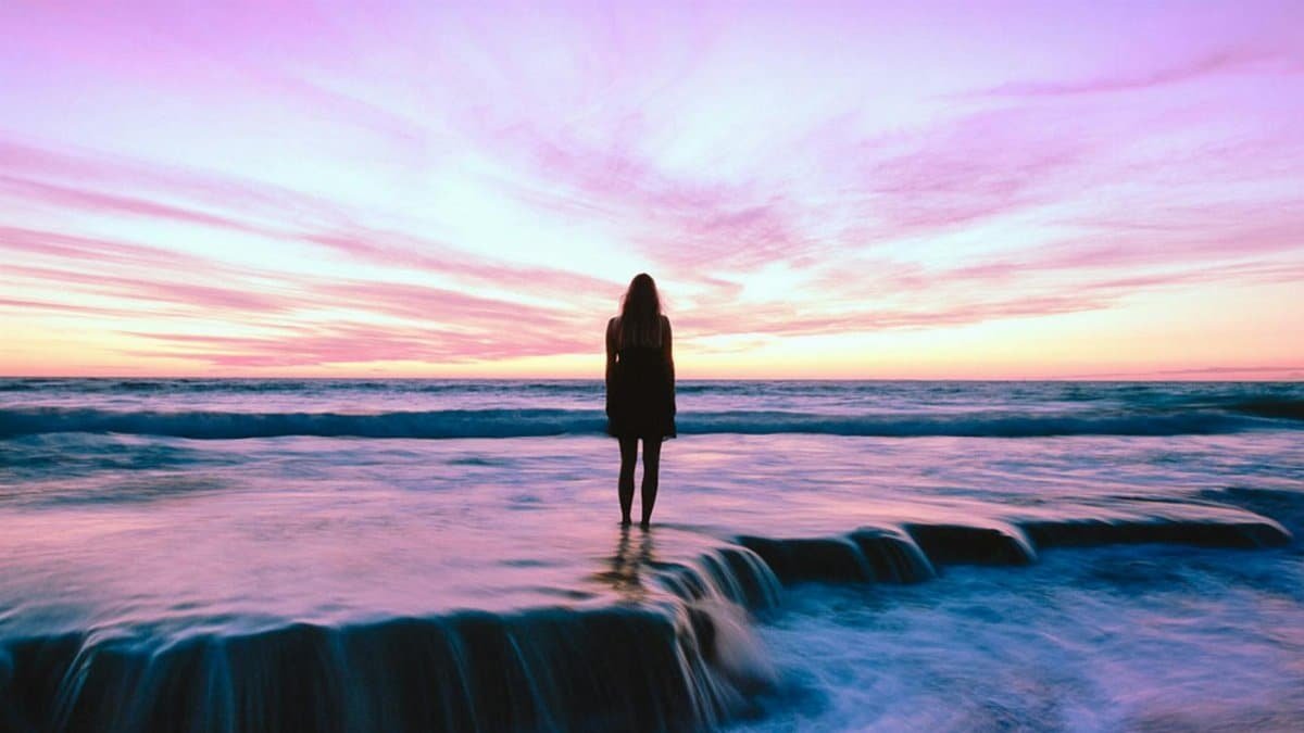 A woman stands alone by the ocean at sunset, embracing nature's beauty.