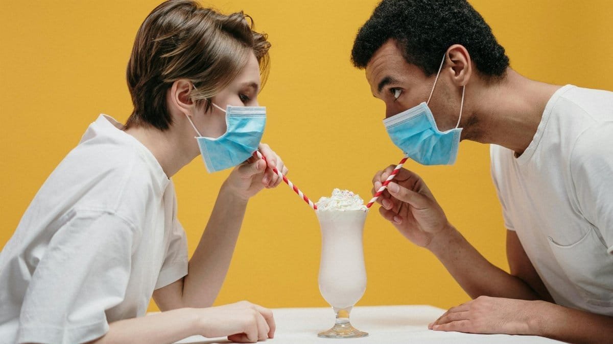 A couple wearing masks humorously shares a milkshake using straws over a white table.