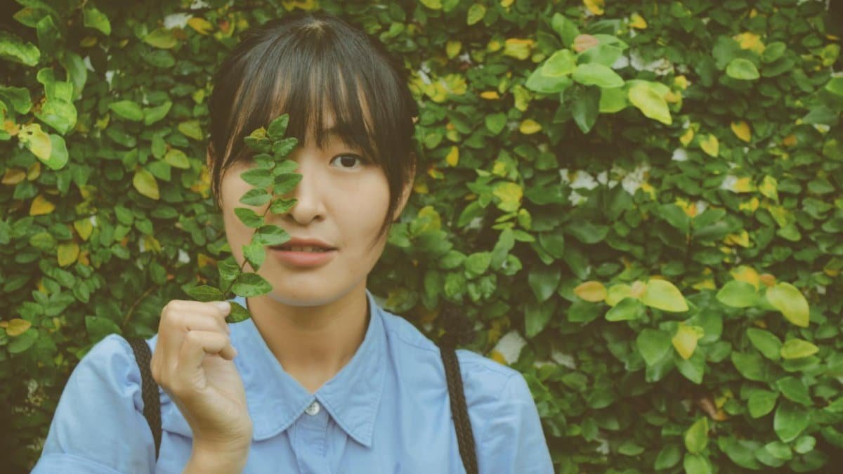 Portrait of young woman holding a green leaf, standing against lush ivy wall background.