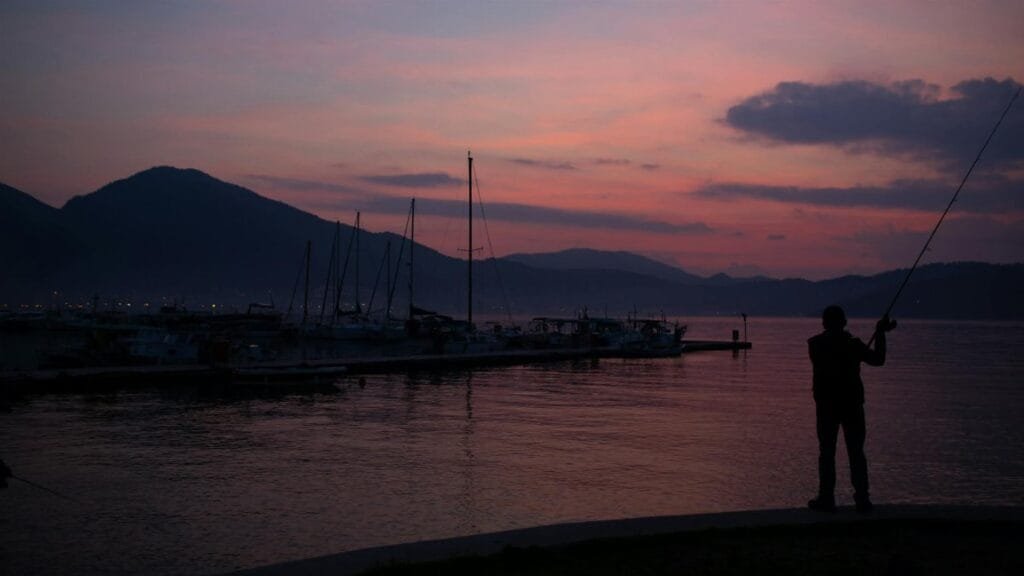 A serene scene of a fisherman silhouetted against a colorful twilight sky by a calm lake.