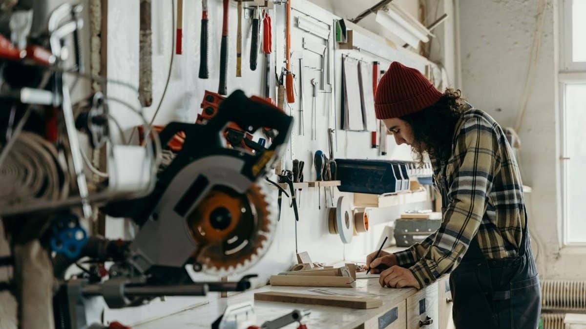 A man wearing a beanie focuses on a project in a tool-filled workshop.