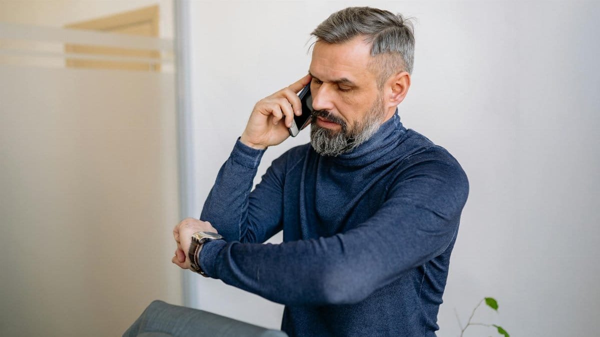 Man with beard speaking on phone while checking the time indoors.