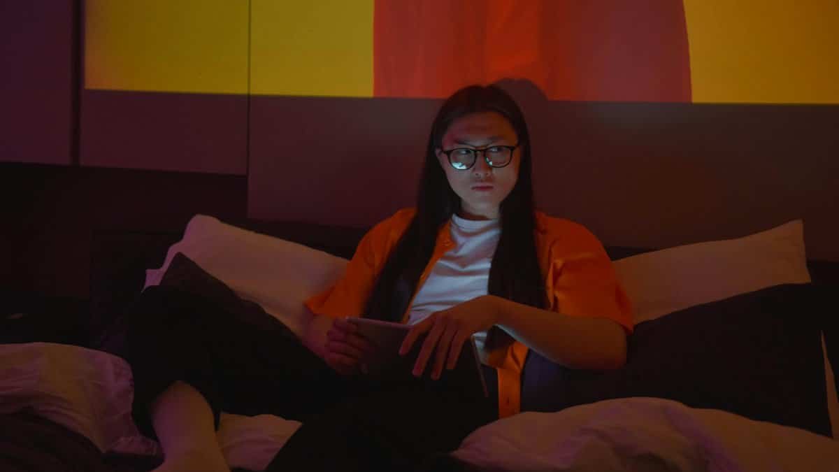 Asian woman with eyeglasses using a tablet, seated indoors with moody lighting.