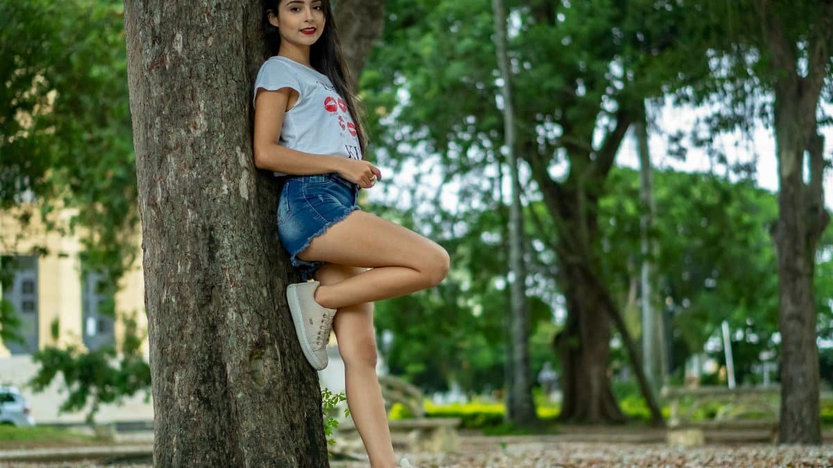 Full body side view of cheerful teen girl in casual clothes leaning on tree trunk in park