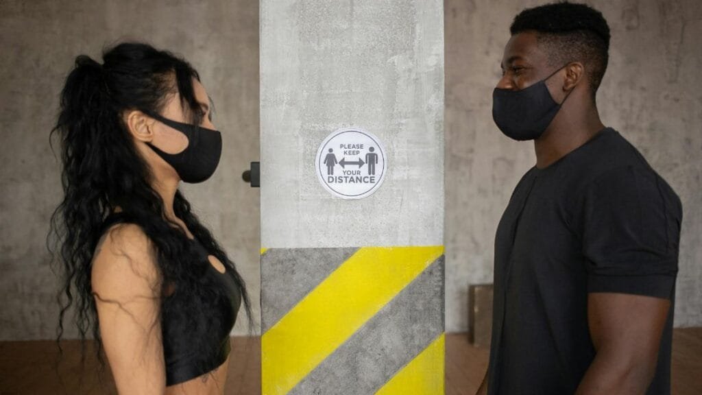 Two people practicing social distancing with masks indoors near a safety sign.