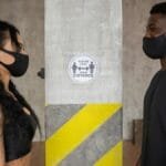 Two people practicing social distancing with masks indoors near a safety sign.