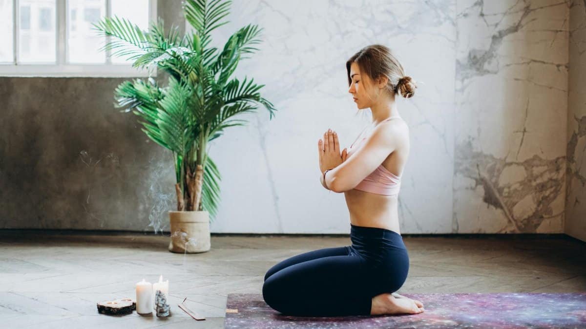 A woman practicing yoga and meditation indoors beside candles and a plant, promoting relaxation and wellness.