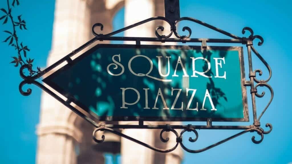 Elegant vintage square piazza sign with ornate iron design, set outdoors under bright sunlight.