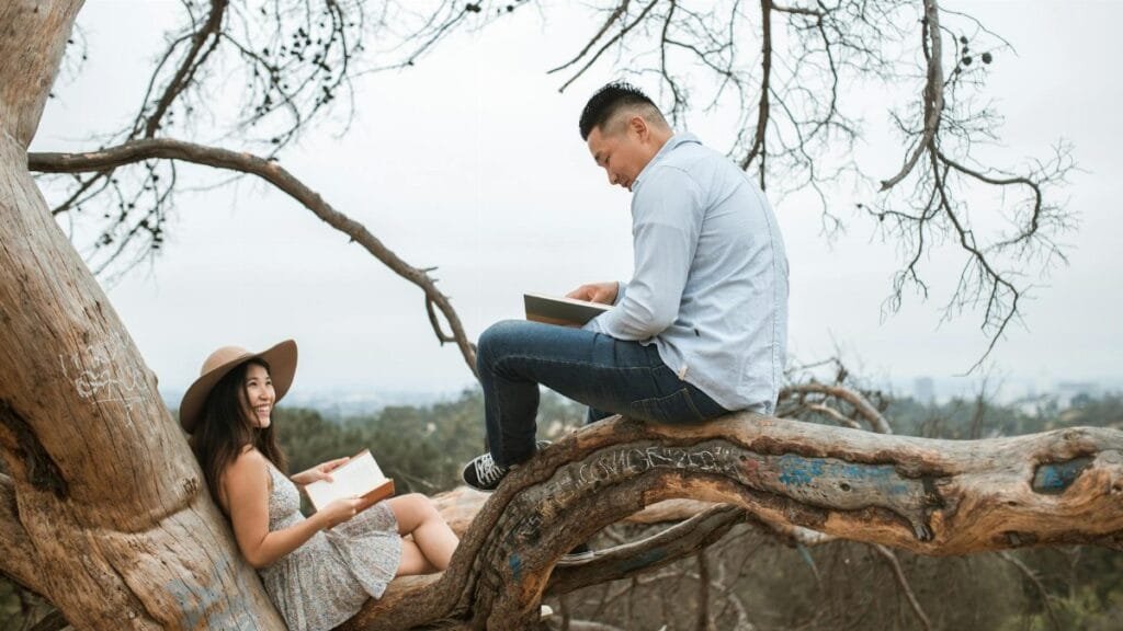 An Asian couple enjoys reading books while sitting in a tree in a park setting.