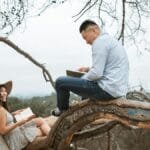 An Asian couple enjoys reading books while sitting in a tree in a park setting.