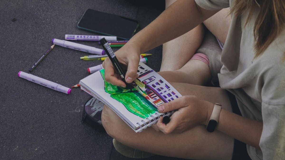 Close-up of a young artist drawing on paper with colored pens, sitting outdoors.