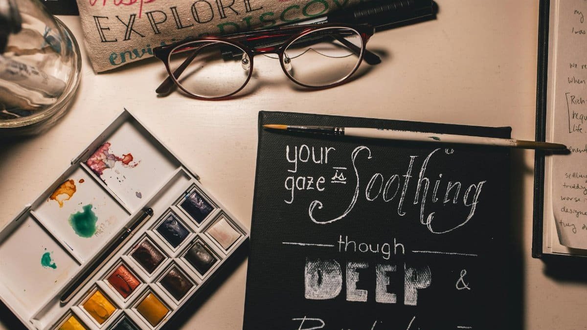 Artistic workspace featuring paints, a canvas with text, and eyeglasses for a creative touch.