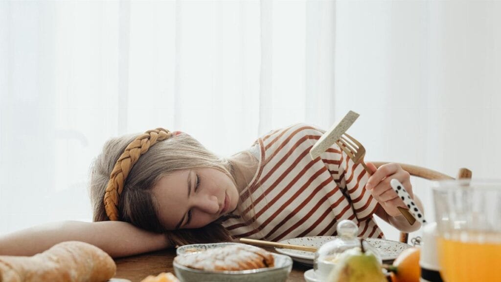 A girl in a striped shirt resting her head on a table surrounded by breakfast foods, holding a fork.