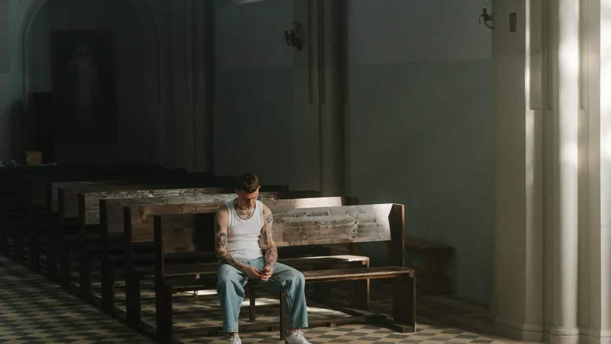A tattooed man contemplates life while sitting alone in a quiet church pew.