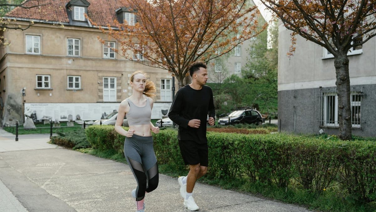 Two adults jogging on a sidewalk in an urban setting surrounded by spring foliage.