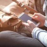 Woman sitting with smartphone and brown bag in sunlight.
