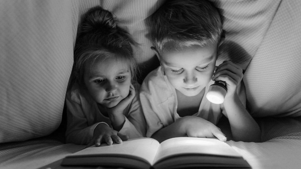 A boy and girl reading with a flashlight under a blanket in grayscale