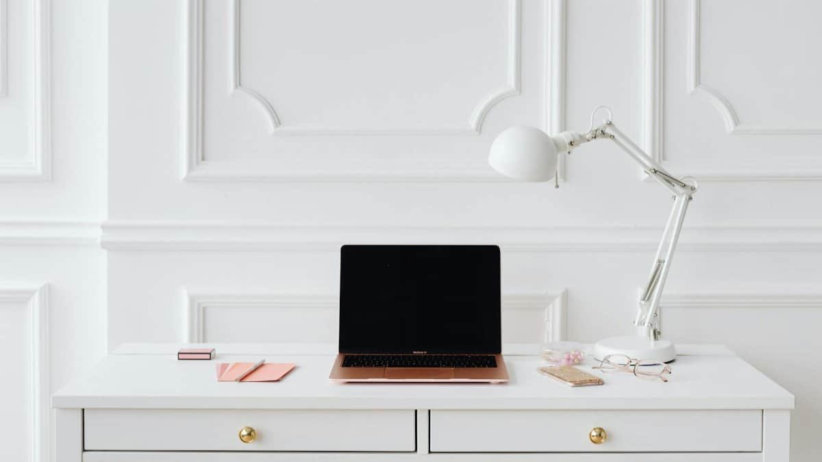A clean and bright workspace with a laptop, desk lamp, and essentials on a white table.