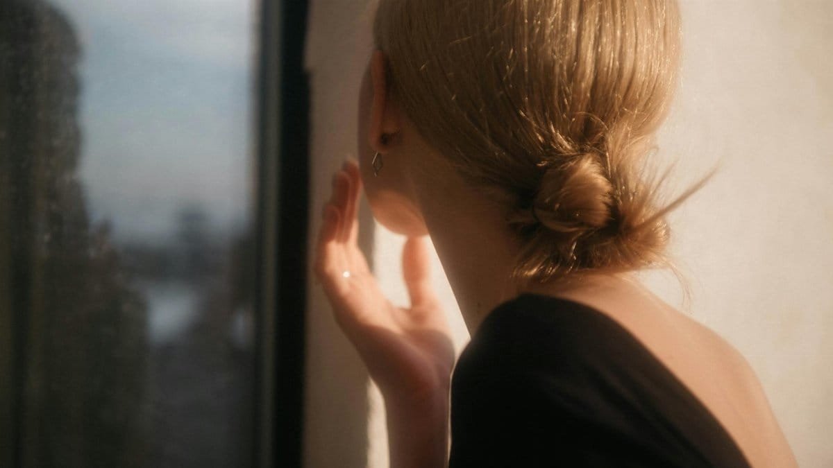 A woman with blonde hair gazes out of a sunlit window, reflecting serenity.