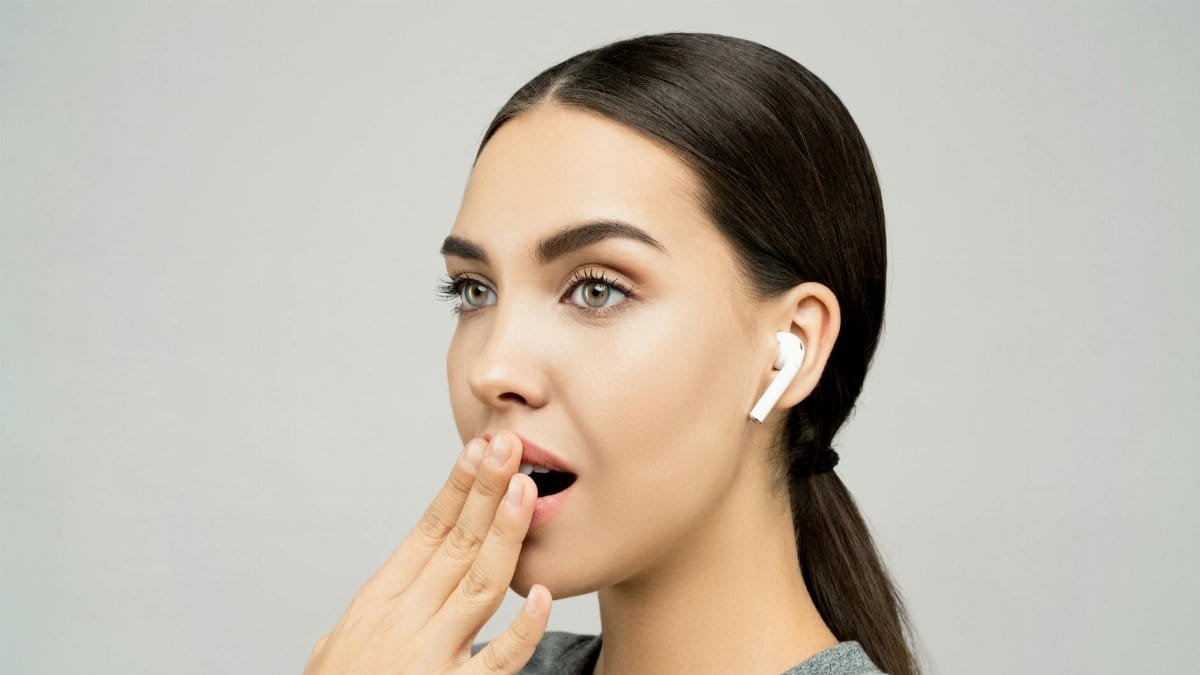 Portrait of a young woman with wireless earbuds, expressing surprise against a gray background.