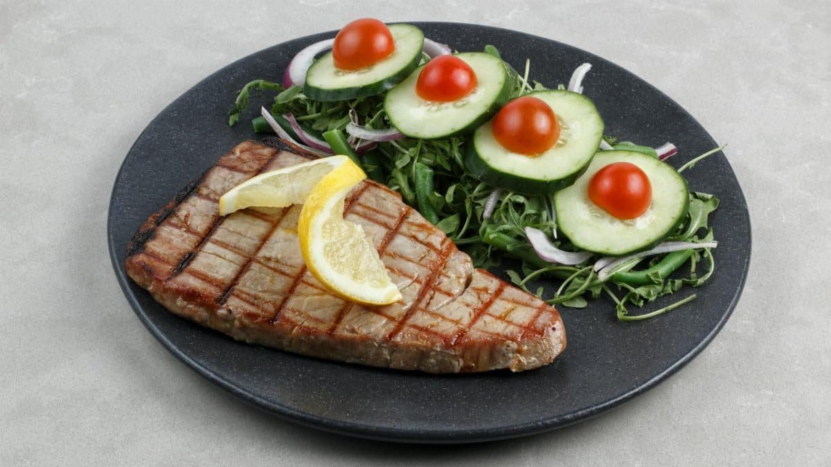 A grilled tuna steak topped with lemon slices served with a fresh cucumber and tomato salad on a dark plate.