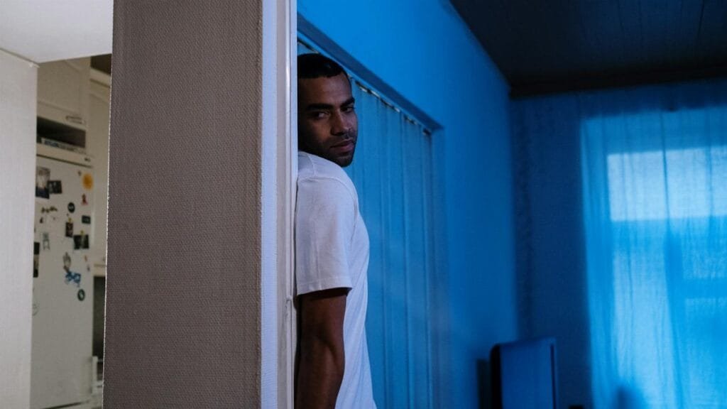 A man contemplatively leans against a wall in a dimly lit indoor setting, under blue light.