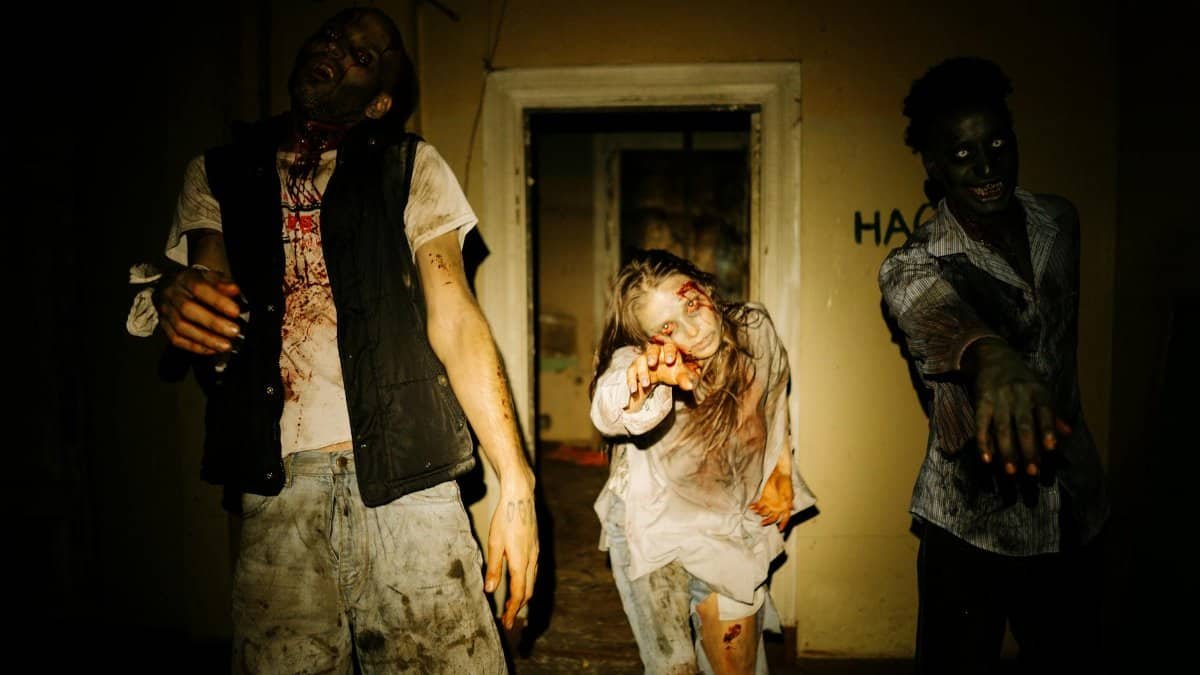 Three costumed zombies outstretch their hands in a dimly lit, abandoned house setting.