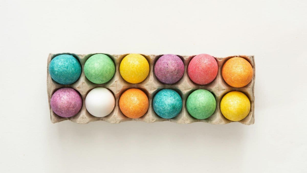Vibrant painted Easter eggs arranged in a tray, showcasing a rainbow of colors on a minimal white background.