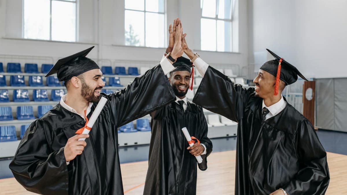 Three graduates in gowns celebrating their success with high fives indoors.