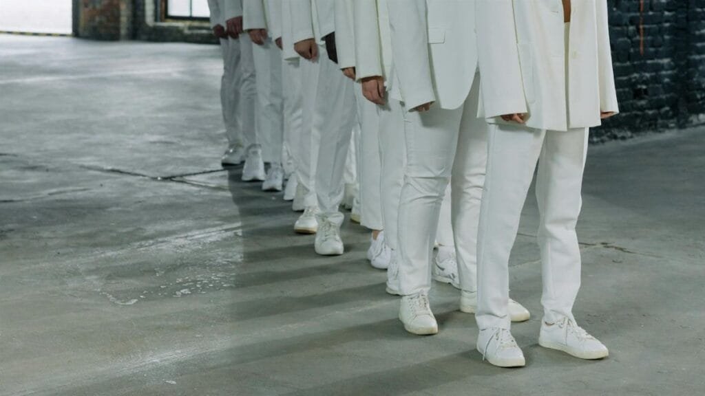 A row of individuals dressed in white suits and sneakers standing in alignment indoors, casting shadows.