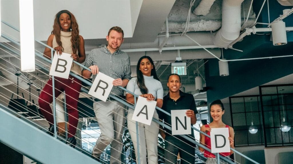 Diverse group of adults smiling and holding 'BRAND' signs on an office staircase.