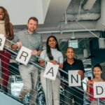Diverse group of adults smiling and holding 'BRAND' signs on an office staircase.