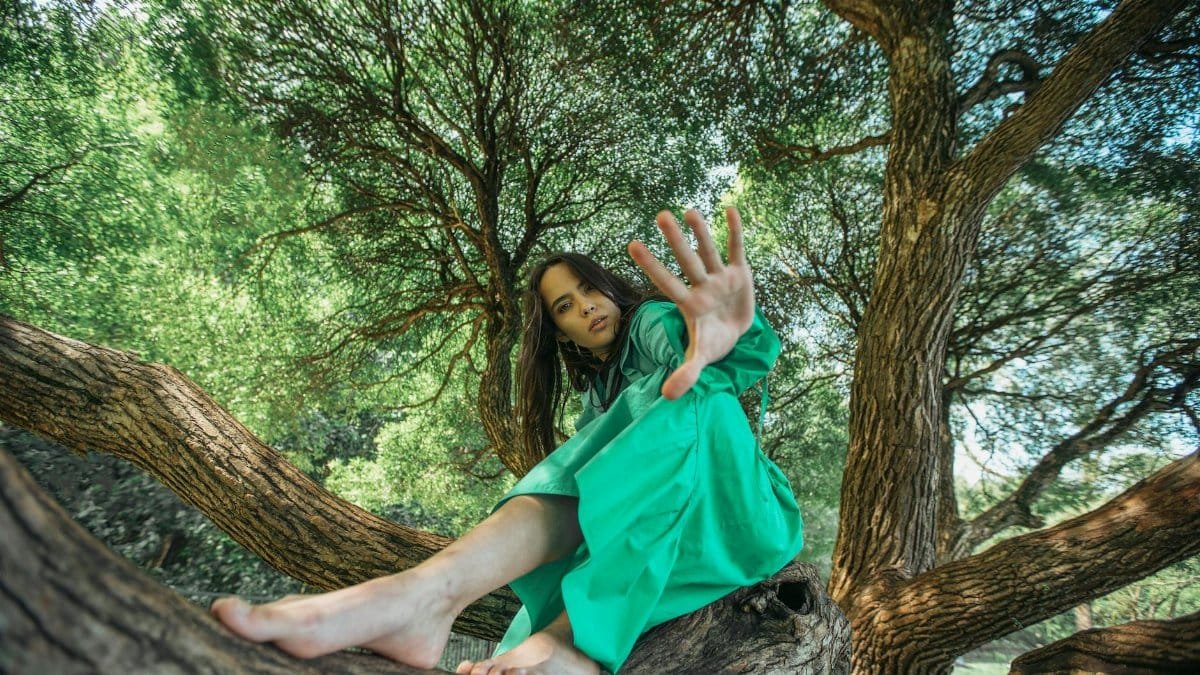 A woman in a green dress sitting on tree branches, surrounded by lush greenery.